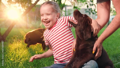 Little girl plays with puppies with her mother in park. Child, mom plays with Labrador puppies on green grass in garden. Joyful child, mom play with their dog. Kind child having fun with dog outdoors photo