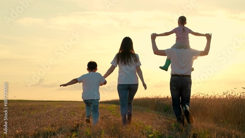 Happy farmers family with children runs through wheat field at sun. Big family, people in nature, holiday. Daughter on shoulders of dad, mom, son, walk hand in hand outdoor. Parental care for children