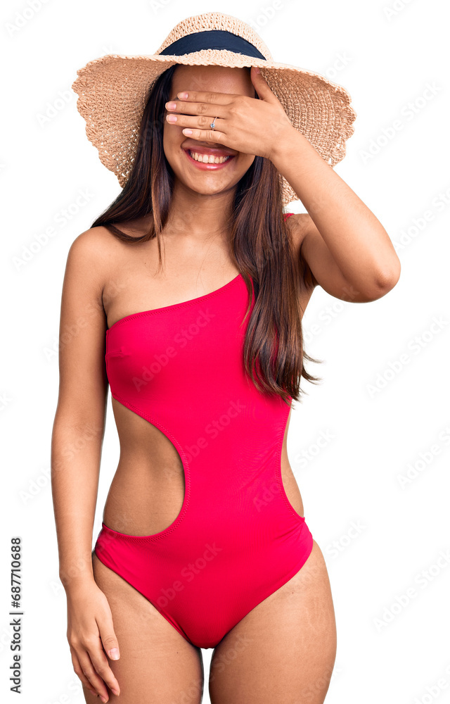 Young beautiful latin girl wearing swimwear and summer hat smiling and laughing with hand on face covering eyes for surprise. blind concept. Young beautiful latin girl wearing swimwear and summer hat smiling and laughing with hand on face covering eyes for surprise. blind concept.