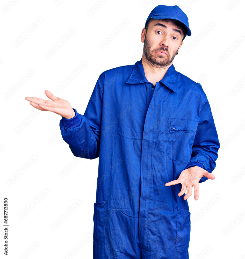 Young hispanic man wearing painter uniform clueless and confused expression with arms and hands raised. doubt concept.