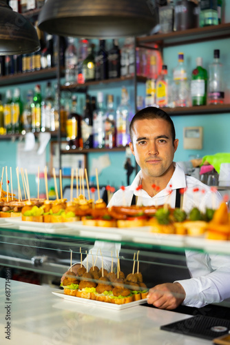 Courageous Latin male barman in apron standing behind the snack bar with pinchos and alcohol. High quality photo