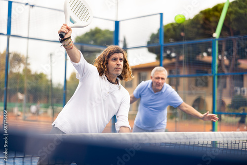 Two confident adult men padel players hitting ball with rackets on hard court in spring