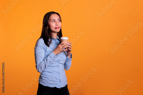 Cheerful model drinking cup of coffee over yellow background, holding hot beverage in front of camera. Positive filipino woman having refreshment drink enjoying cappucino while posing in studio