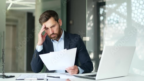 Confused puzzled businessman in a formal suit having difficulty with paper work sitting at workplace in business office. Frustrated financier reviewing documents, unhappy with bad financial results photo