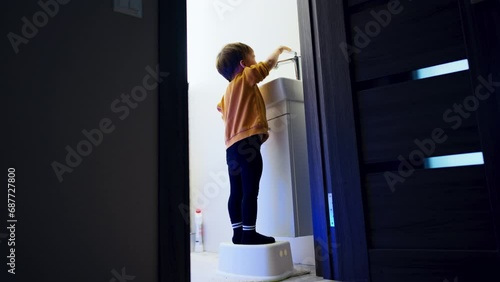Toddler in sweater and tights stands on the prop in the bathroom. Kid looks into the sink watching the water flow. Low angle view. photo