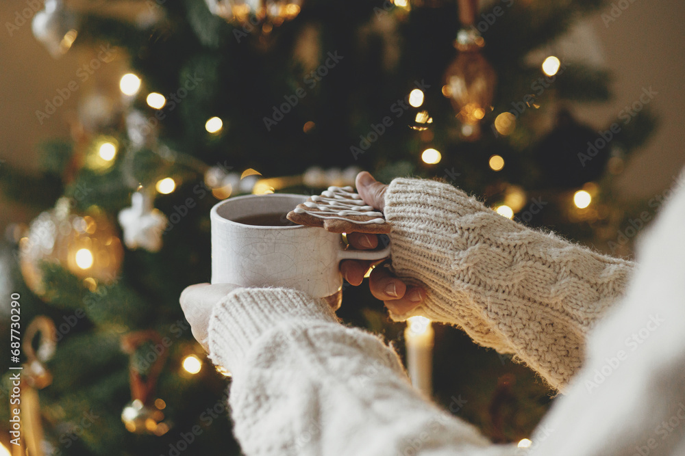 Hands in sweater holding warm cup of tea with christmas gingerbread cookie against festive christmas tree with golden lights bokeh. Merry Christmas! Atmospheric holiday time, winter hygge
