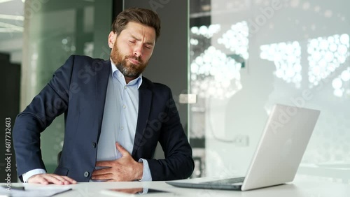 Businessman in a formal suit feels stomach pain while working on a laptop while sitting at workplace in business office. Sick worker has heartburn, gastritis or poisoning. Male suffers from spasms photo