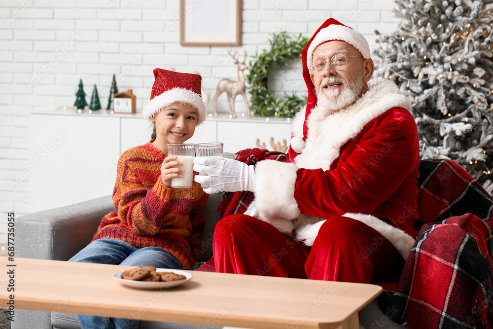 Fototapeta premium Santa Claus and cute little girl drinking milk with cookies on sofa in decorated room for Christmas