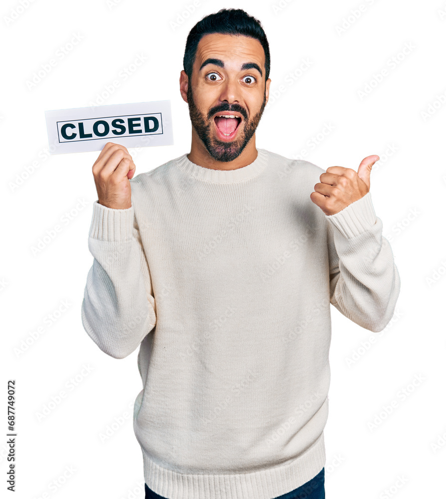Young hispanic man with beard holding closed banner pointing thumb up to the side smiling happy with open mouth Young hispanic man with beard holding closed banner pointing thumb up to the side smiling happy with open mouth