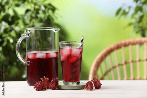 Refreshing hibiscus tea with ice cubes and roselle flowers on white wooden table against blurred green background. Space for text photo