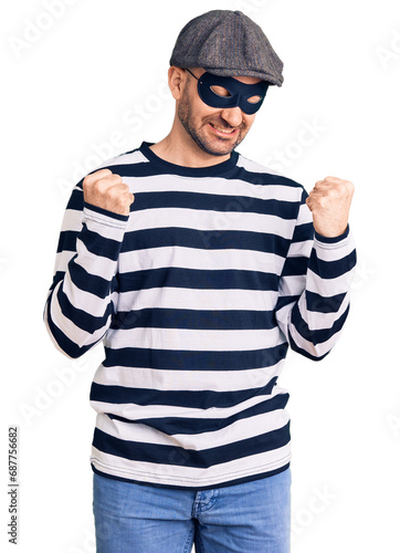Young handsome man wearing burglar mask very happy and excited doing winner gesture with arms raised, smiling and screaming for success. celebration concept.