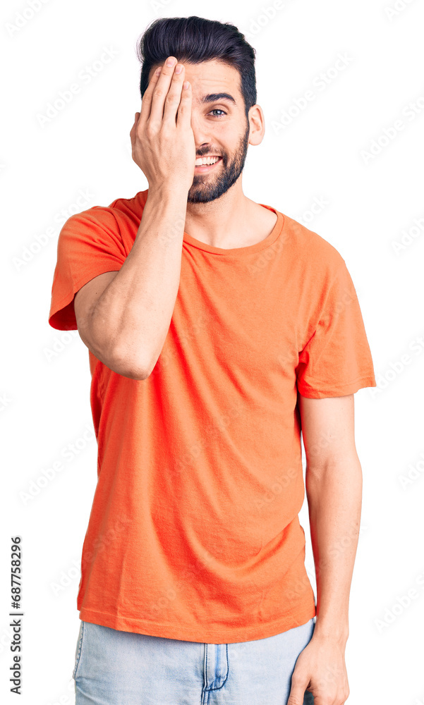 Young handsome man with beard wearing casual t-shirt covering one eye with hand, confident smile on face and surprise emotion. Young handsome man with beard wearing casual t-shirt covering one eye with hand, confident smile on face and surprise emotion.