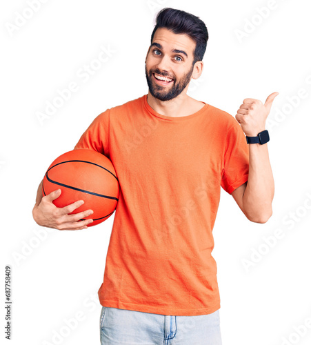 Young handsome man with beard hoilding basketball ball pointing thumb up to the side smiling happy with open mouth © Krakenimages.com