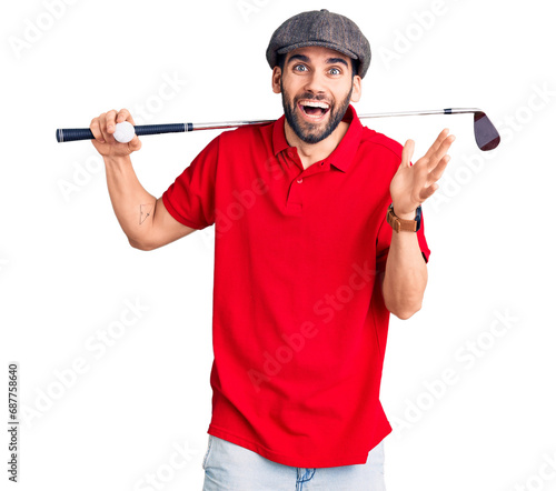 Young handsome man with beard playing golf holding club and ball celebrating victory with happy smile and winner expression with raised hands