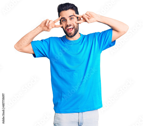 Young handsome man with beard wearing casual t-shirt doing peace symbol with fingers over face, smiling cheerful showing victory