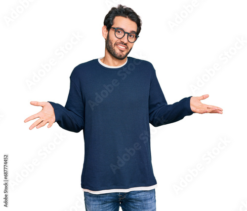 Young hispanic man wearing casual clothes and glasses clueless and confused expression with arms and hands raised. doubt concept.