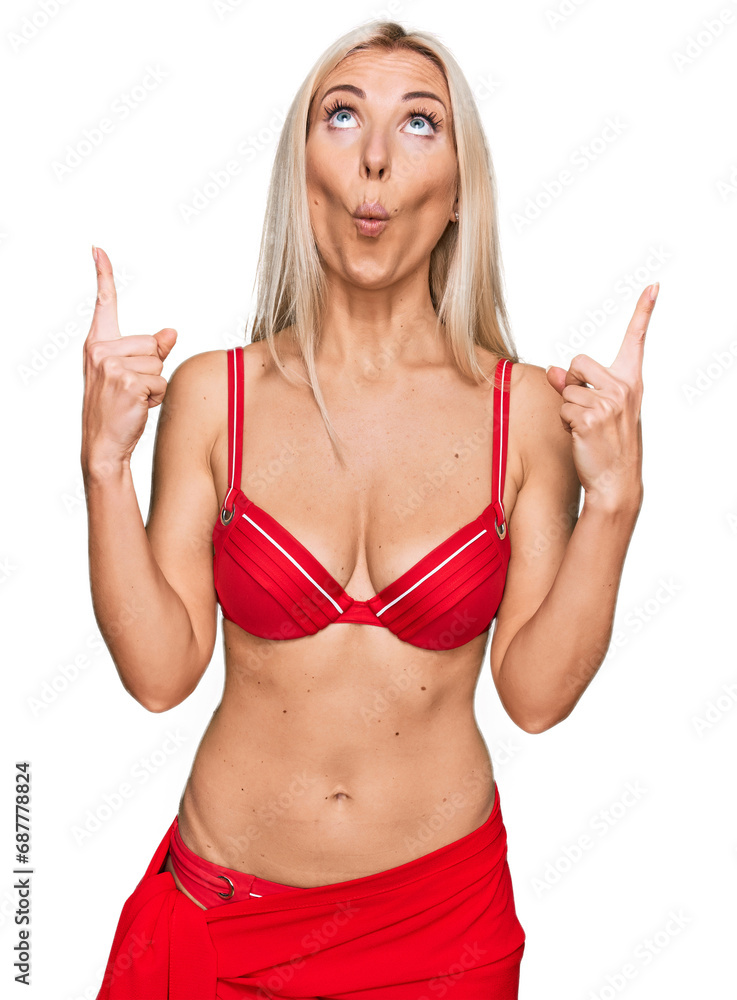 Young blonde woman wearing bikini amazed and surprised looking up and pointing with fingers and raised arms. Young blonde woman wearing bikini amazed and surprised looking up and pointing with fingers and raised arms.
