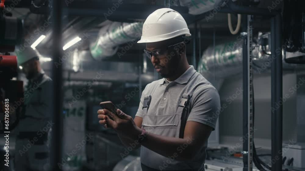 African American engineer uses mobile phone to inspect pipeline system on plant. Professional male worker in safety uniform and protective hard hat works on modern factory or industrial facility. African American engineer uses mobile phone to inspect pipeline system on plant. Professional male worker in safety uniform and protective hard hat works on modern factory or industrial facility.