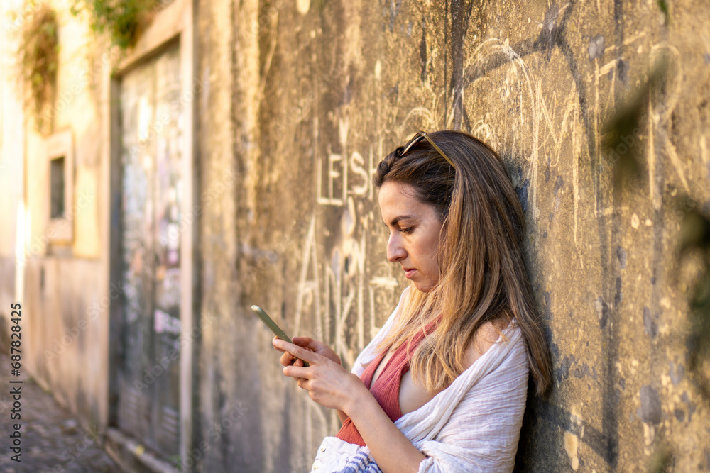 Obraz premium Engrossed in Technology: A Side-View, Horizontal Shot of an Unidentified Individual Leaning Against a Wall, Engaged in Texting on Their Phone, Under the Warm Sunlight of the Day