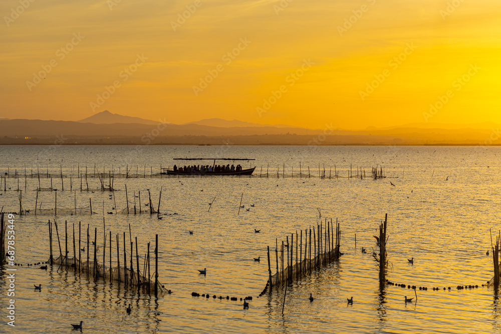 A boat with tourists and fishermen on a lake with fish nets and sunny path against the backdrop of an orange sunset
