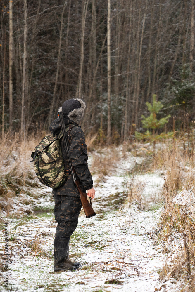 Male hunter in camouflage and with backpack, armed with a rifle, walks through the snowy winter forest