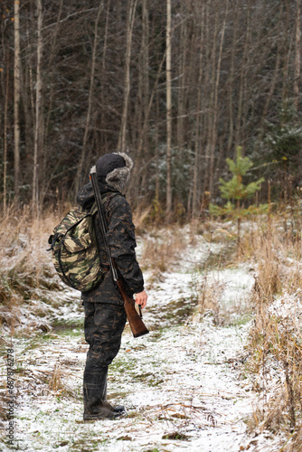 Male hunter in camouflage and with backpack, armed with a rifle, walks through the snowy winter forest