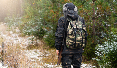 Male hunter in camouflage and with backpack, armed with a rifle, walks through the snowy winter forest