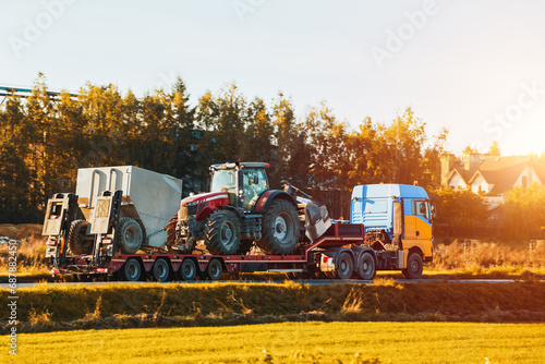A heavy truck with a flatbed trailer transports a big tractor on the road. The photo shows the industrial machinery and equipment used for farming and agriculture. The truck is driving in the evening photo