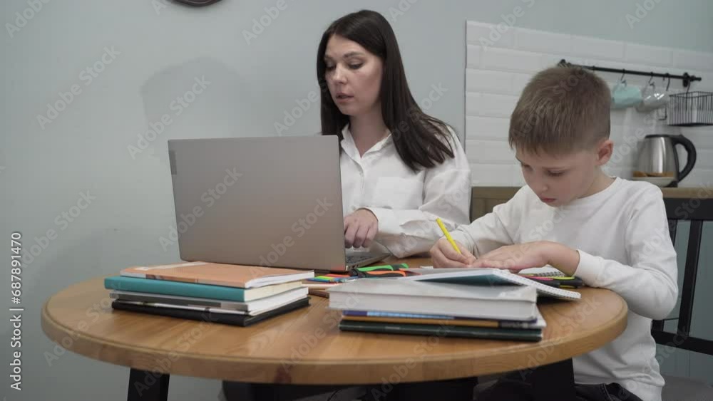 Medium shot boy drawing while his mother is working at a computer sitting together at a table in the kitchen. Happy family. Medium shot boy drawing while his mother is working at a computer sitting together at a table in the kitchen. Happy family.