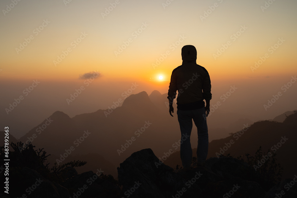 Silhouette of a hiker man on a rock pedestal with hands up. Beautiful orange sunset. Independent hiking travel, success.