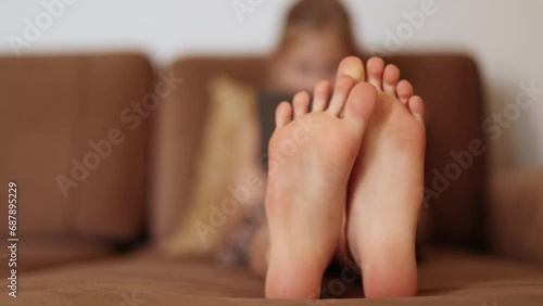 Close of of little girl's bare feet as she sits on a couch at home and reads a book or plays in a tablet, leisure time at home concept photo