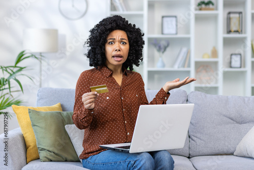 Portrait of a worried young African-American woman sitting on the sofa at home with laptop, holding a credit card, and waving her hands anxiously