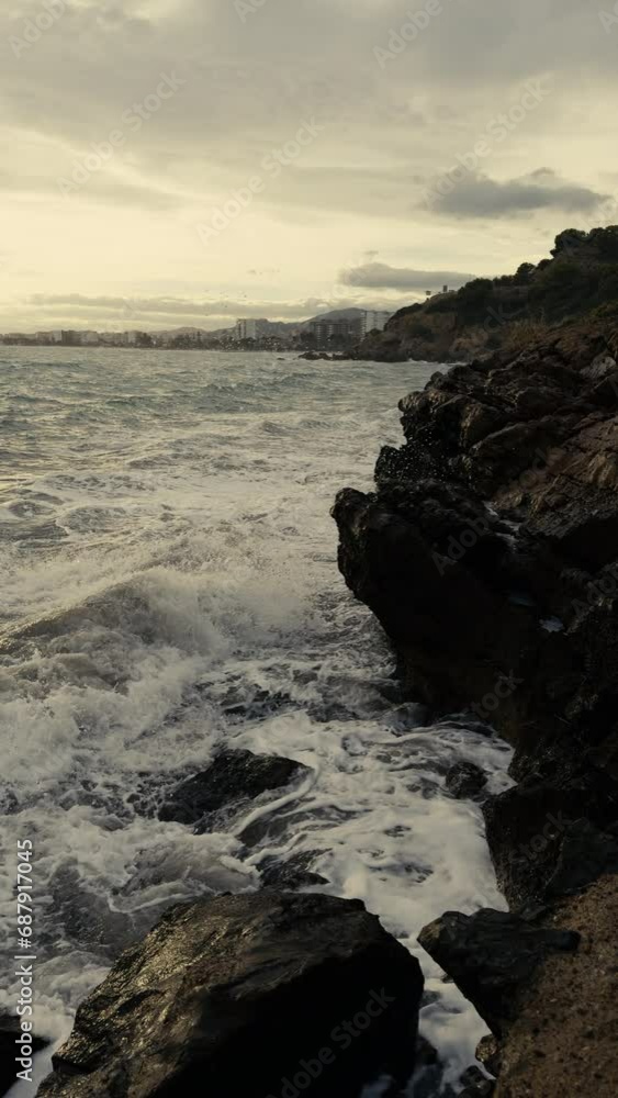 Storm at sea in winter in Benicassim. Waves crash against the rocks on a winter evening. On the horizon there is a city against the backdrop of mountains. Spain. Province of Castellon. Slow motion Storm at sea in winter in Benicassim. Waves crash against the rocks on a winter evening. On the horizon there is a city against the backdrop of mountains. Spain. Province of Castellon. Slow motion