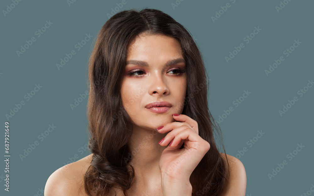 Pensive young model woman with wavy dark brown hairstyle and fresh clean shiny skin on blue studio background, fashion portrait. Skin care, hair care and cosmetology concept Pensive young model woman with wavy dark brown hairstyle and fresh clean shiny skin on blue studio background, fashion portrait. Skin care, hair care and cosmetology concept