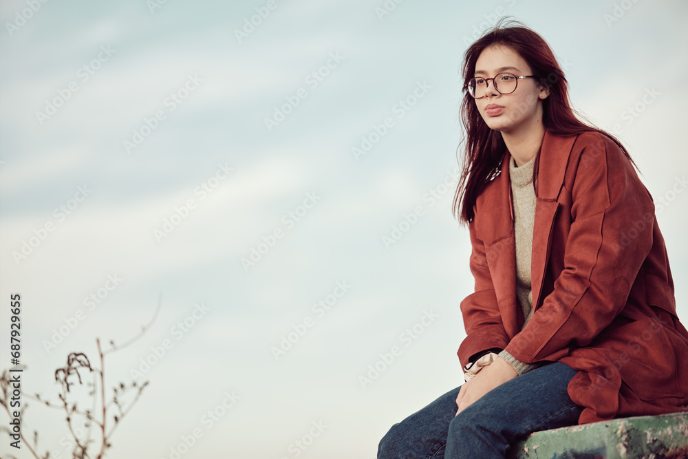 Pensive calm teenage girl in glasses with long red hair in red coat sits on concrete podium and looks away, against the background of sky . Pensive calm teenage girl in glasses with long red hair in red coat sits on concrete podium and looks away, against the background of sky .
