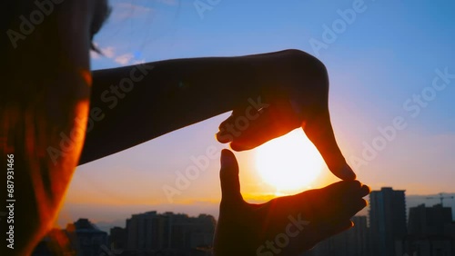 Close up view: silhouette of woman hands are doing finger frame and looking at the sunset, sunrise sky over the city with sun lens flares. Imagination, creativity, gesture and composition concept photo