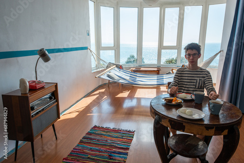 Handsome teen boy eating in vintage room with old fashioned armchair, table with food, telephone, standart lamp, doormat, chairs, hammock and sea view. Interior of 20th century, nostalgia photo