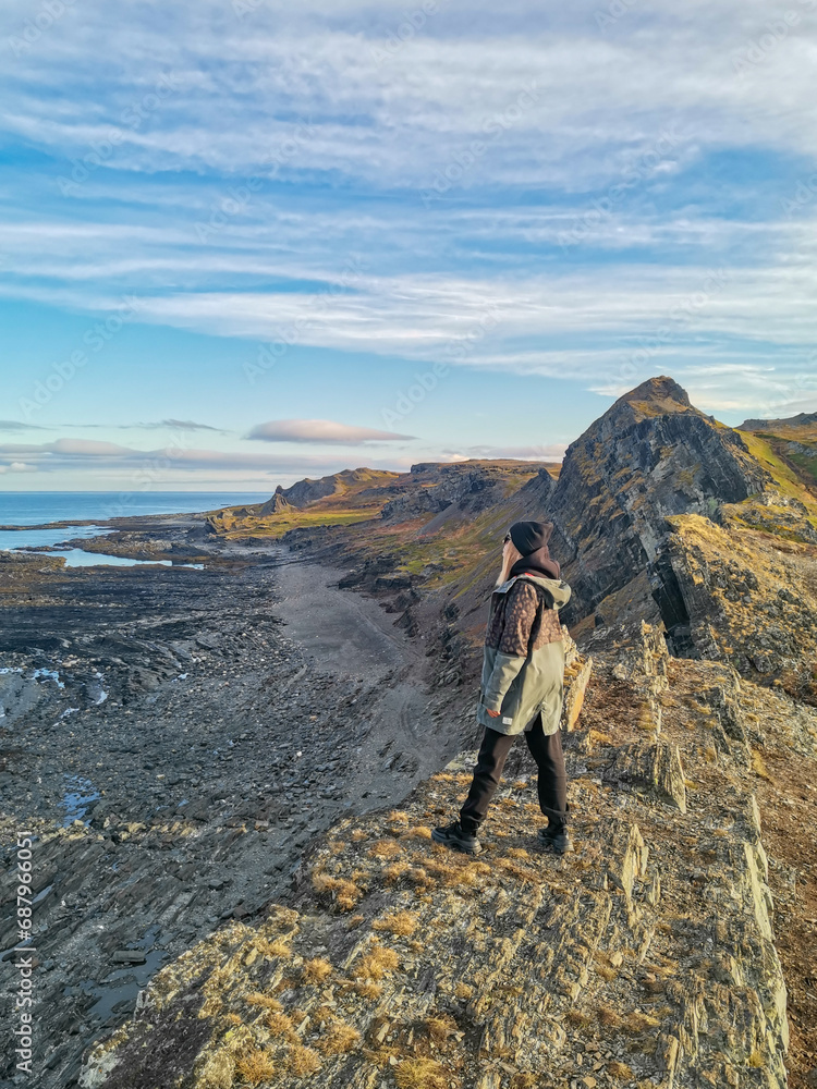 Fototapeta premium A woman on the background of the rocky coast of the Barents Sea. Beautiful view of the cliffs and the coast of the Rybachy and Sredny peninsulas. The harsh beauty of the north.