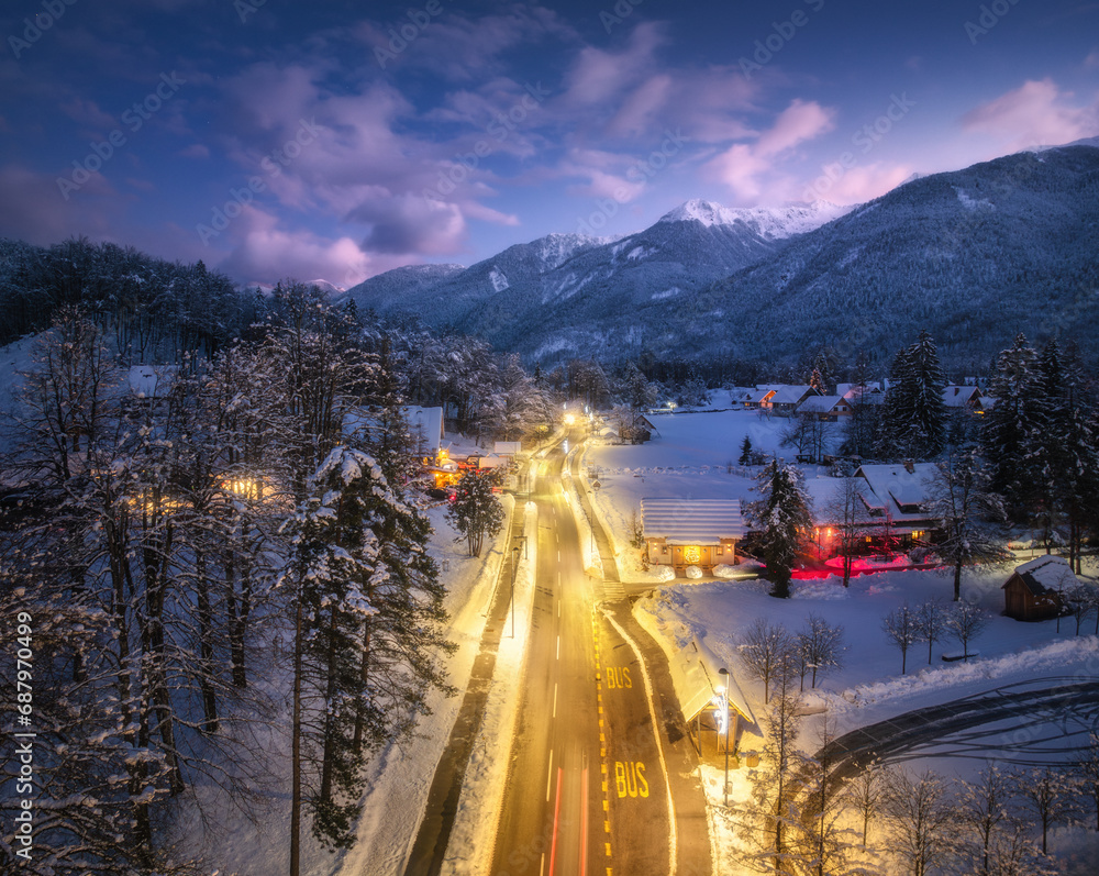 Aerial view of road, snowy mountains, street lights, forest in snow, purple sky at night in winter. Top drone view of alpine village, houses, illumination, highway, cars, pine trees at dusk. Slovenia
