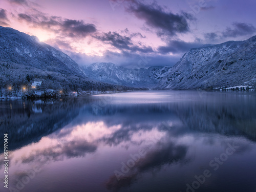 Aerial view of lake, snowy alpine mountains, pine trees in snow, reflection in water, purple sky with clouds at winter night. Nature. Top drone view of beautiful Bohinj lake in Slovenia at sunset