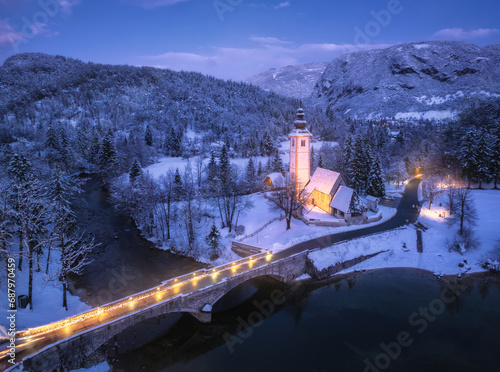 Aerial view of snowy mountains, trees, church, bridge, river, road, street lights, houses, purple sky with clouds at cold winter night. Top drone view of Bohinj lake in Slovenia at twilight. Landscape