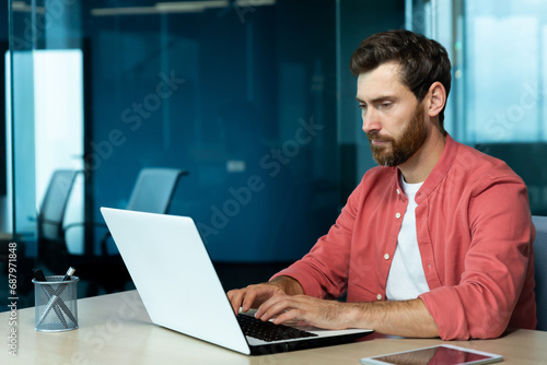 Serious concentrated thinking businessman working with laptop inside office at workplace, man typing on keyboard, preparing online presentation and financial accounting report.