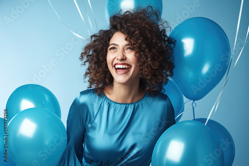 Cheerful curly haired woman dressed in blue clothes, laughing around inflated helium balloons over over background. © Bojan
