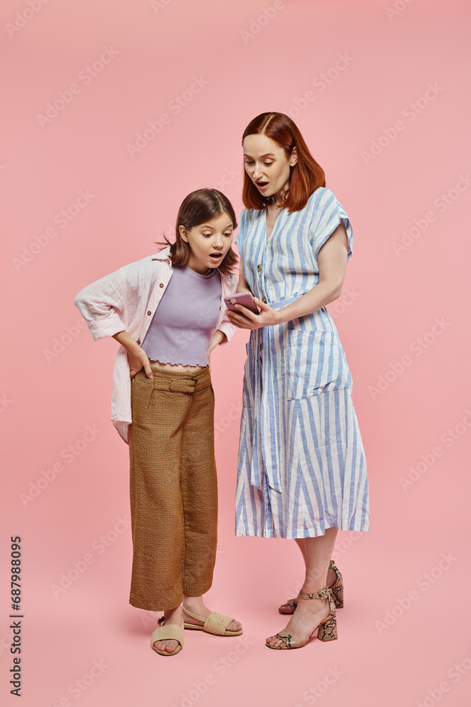 amazed mother and teenage daughter looking at mobile phone while standing on pink backdrop amazed mother and teenage daughter looking at mobile phone while standing on pink backdrop