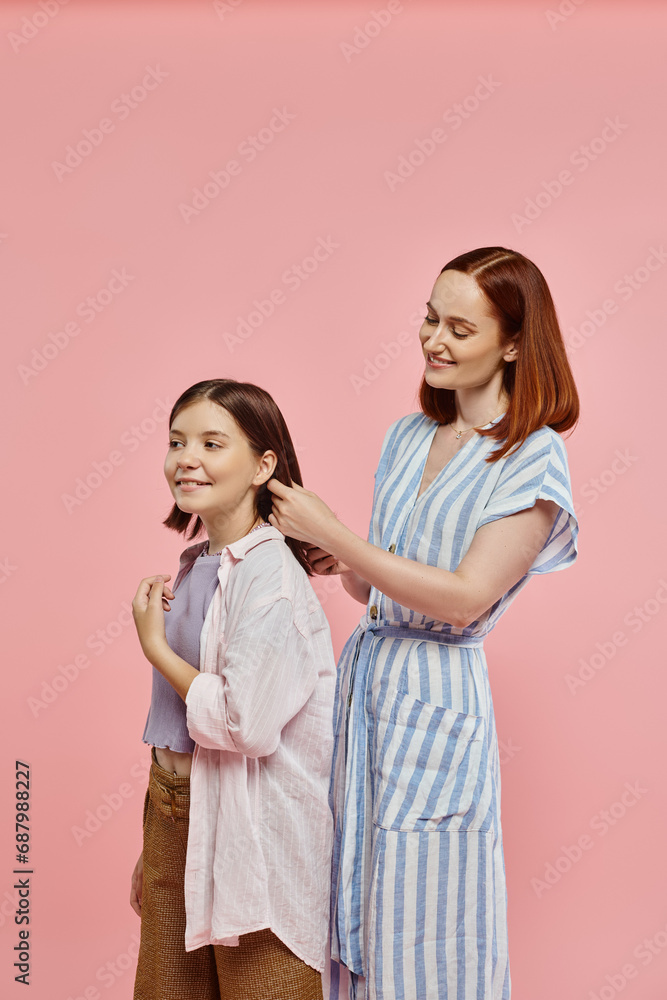 smiling mother braiding hair of happy teenage daughter on pink backdrop in studio, care and love smiling mother braiding hair of happy teenage daughter on pink backdrop in studio, care and love