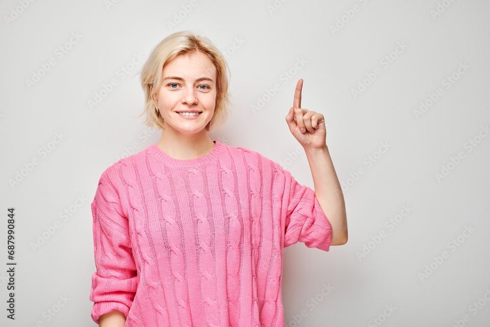 Young blond woman in pink sweater pointing up finger, demonstrating empty space for product or text isolated on white studio background. Young blond woman in pink sweater pointing up finger, demonstrating empty space for product or text isolated on white studio background.
