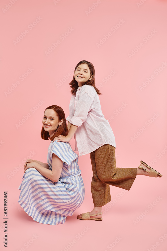 cheerful stylish woman sitting on haunches near teen daughter posing on pink backdrop, fun and joy