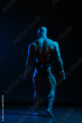 Athletic man demonstrates muscles in the light of a blue light filter on a dark background, view from the back. © ksi