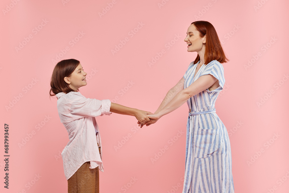 side view of joyful and trendy woman with teenage daughter holding hands on pink, happy family