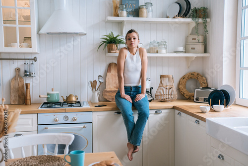 Unhappy brunette girl in casual clothes sitting at kitchen feels loneliness.Disappointed housewife bored at home after divorce. Domestic leisure, sad people. photo